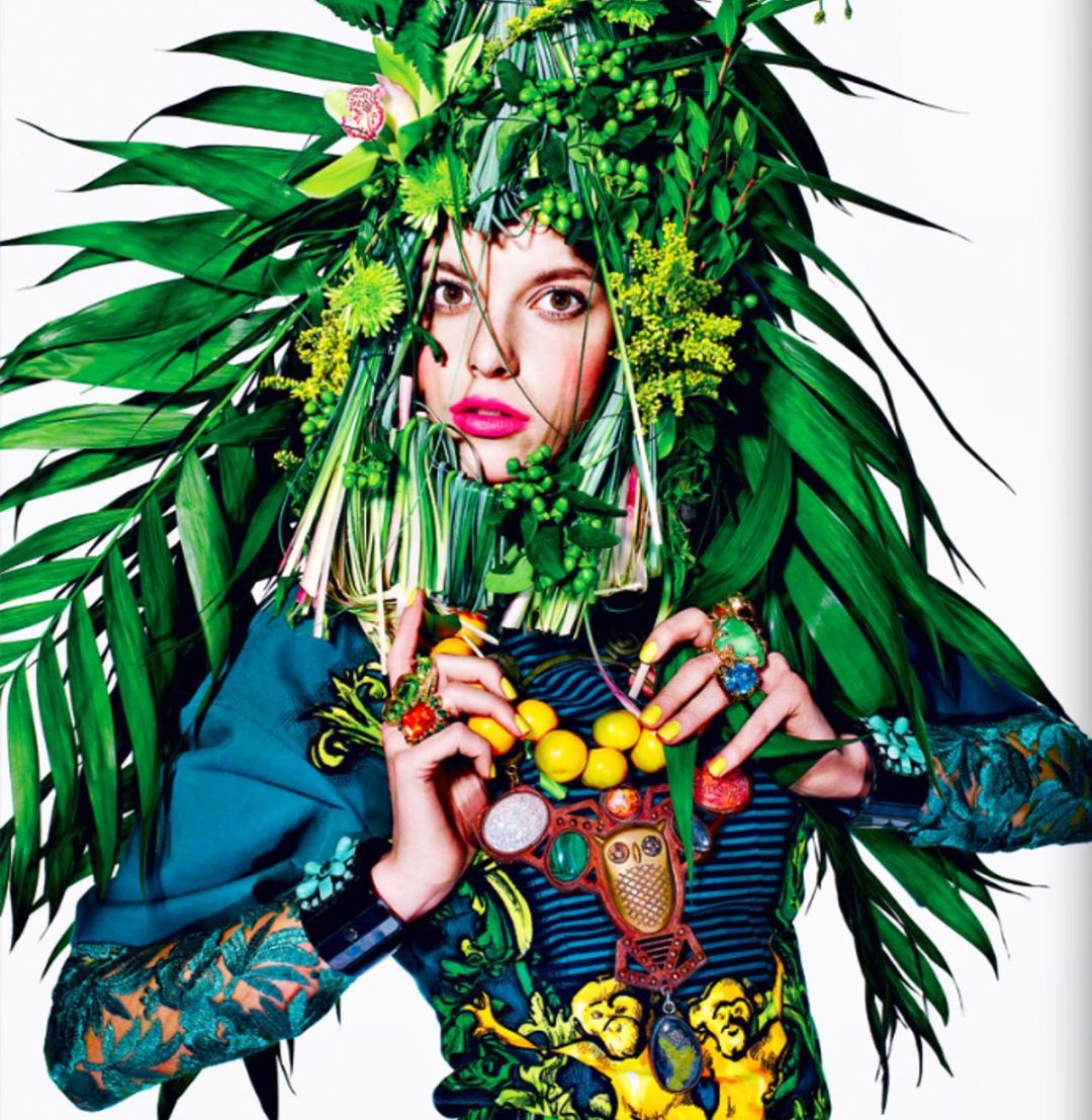 A woman with bright pink lips and a decorative headdress of green leaves and yellow flowers poses for a portrait with colorful jewelry.