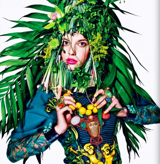 A woman with bright pink lips and a decorative headdress of green leaves and yellow flowers poses for a portrait with colorful jewelry.