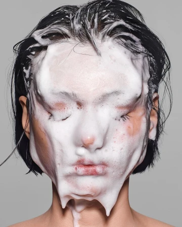 Close-up portrait of a model with dark hair and closed eyes, face covered in white soapy foam against a solid grey studio backdrop.