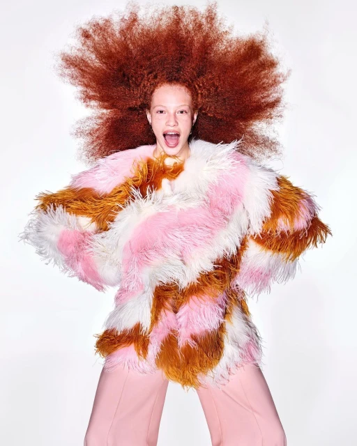 Portrait of a young woman with curly, copper-red hair wearing a colourful faux fur coat and pink pants against a bright white background.