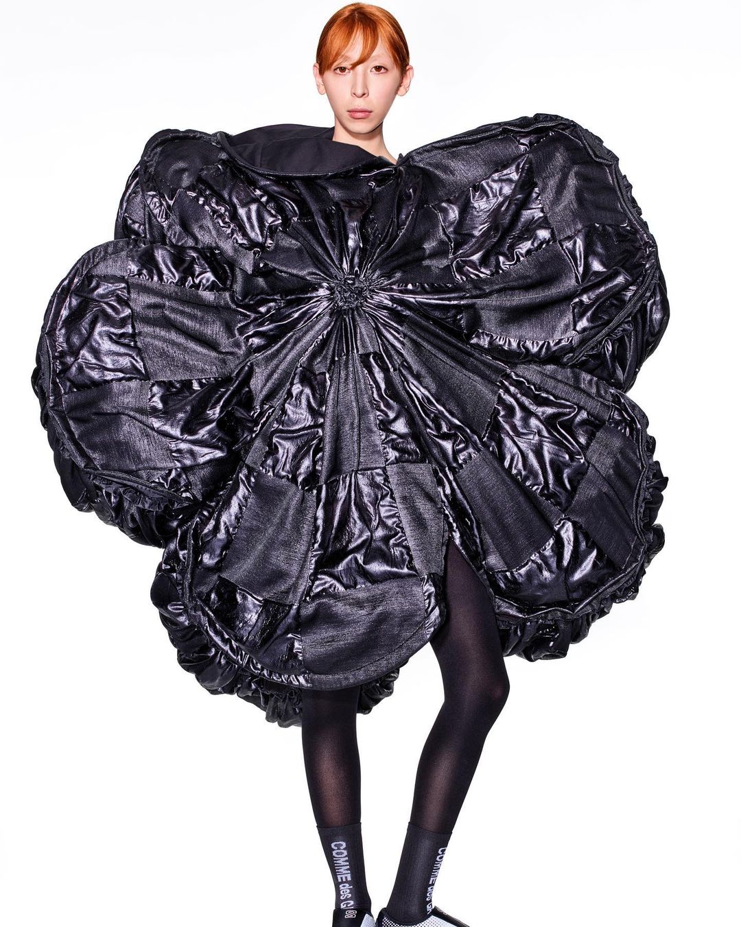 Fashion model poses in an oversized, black, shiny quilted dress resembling a flower against a white background in a full shot.