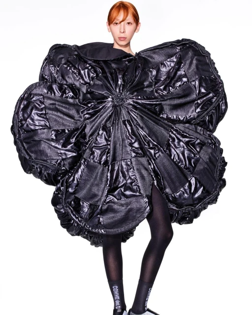 Fashion model poses in an oversized, black, shiny quilted dress resembling a flower against a white background in a full shot.
