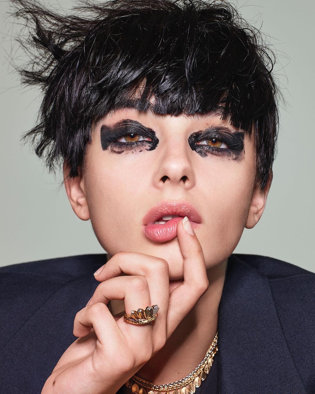 Close-up portrait of a woman with dark hair and dramatic eye makeup wearing a black blazer and jewelry against a light background.