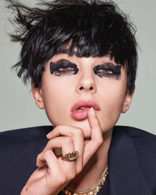 Close-up portrait of a woman with dark hair and dramatic eye makeup wearing a black blazer and jewelry against a light background.