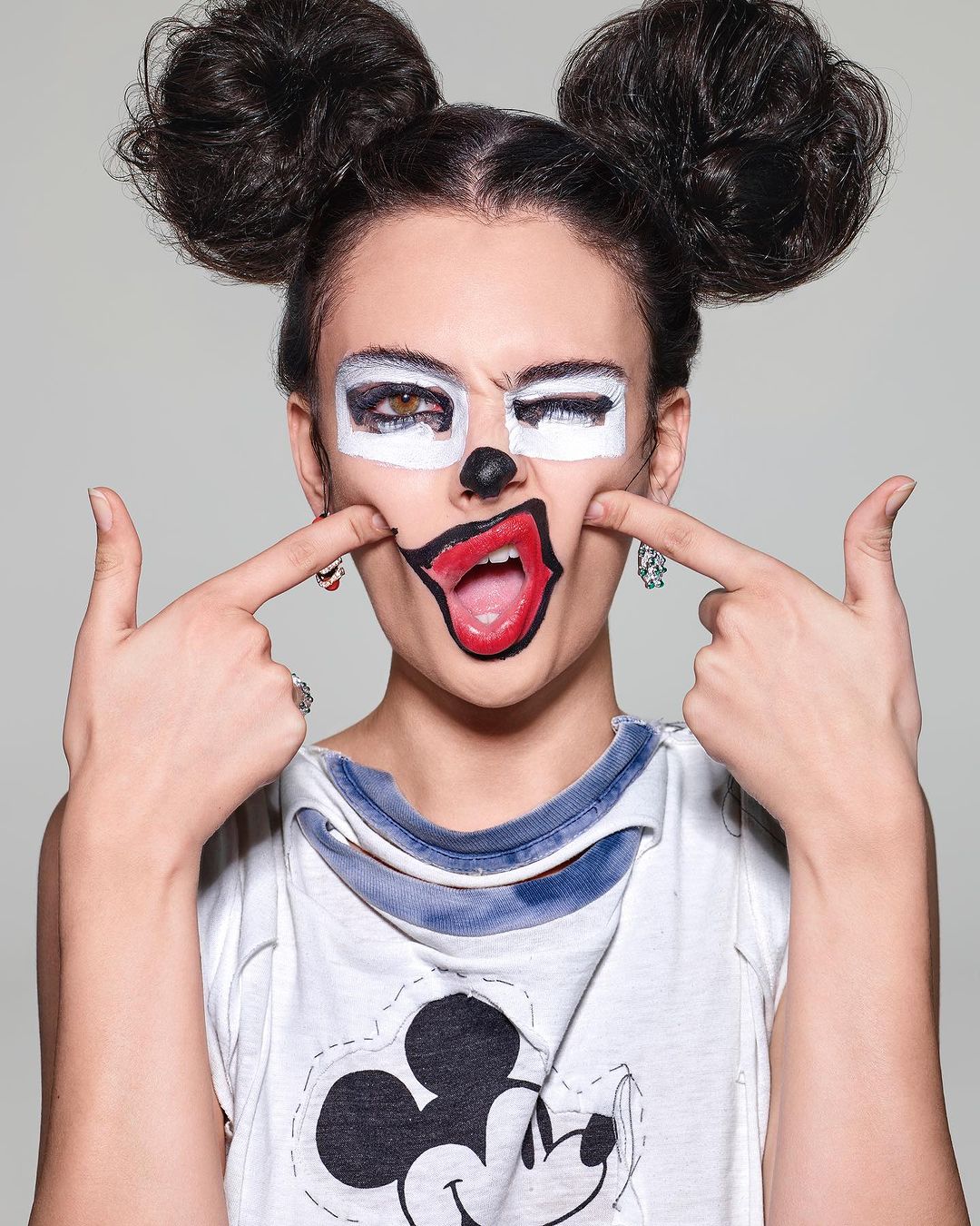 Woman with painted face in cartoon style, wearing Mickey Mouse shirt against a grey background in a medium studio shot.