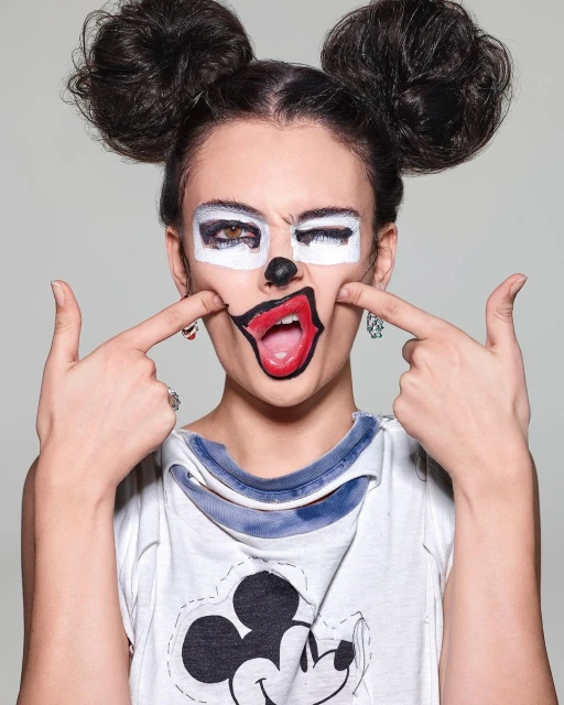 Woman with painted face in cartoon style, wearing Mickey Mouse shirt against a grey background in a medium studio shot.
