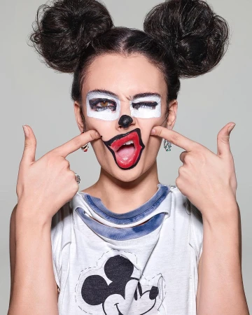 Woman with painted face in cartoon style, wearing Mickey Mouse shirt against a grey background in a medium studio shot.