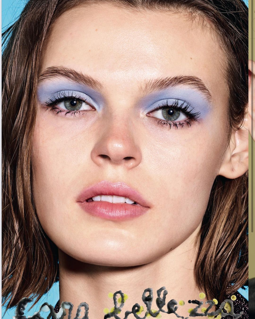 Close-up studio portrait of a young woman with light brown hair, wearing blue eyeshadow and light pink lipstick, beauty product photography.