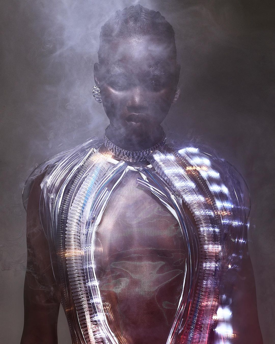 Close-up studio shot of a black female model wearing a silver necklace and futuristic shiny silver apparel with wisps of smoke around her face.
