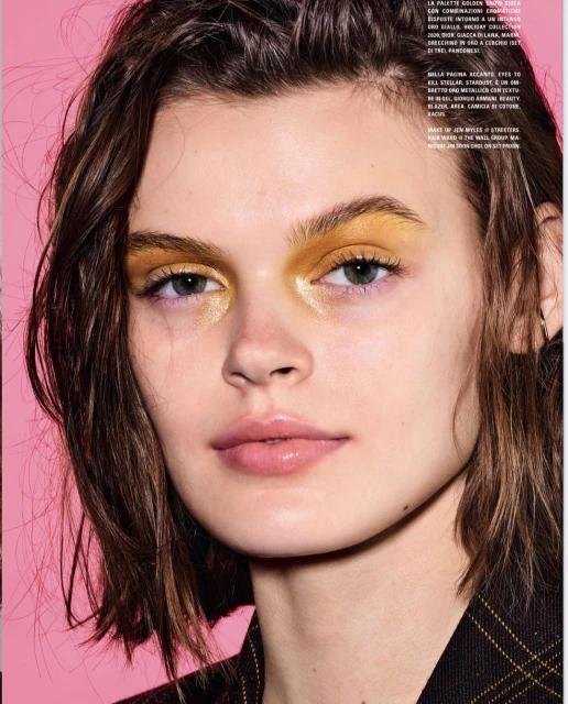 Close-up beauty shot of a young woman with gold eyeshadow and glossy lips against a pink background, showcasing a beauty editorial look.