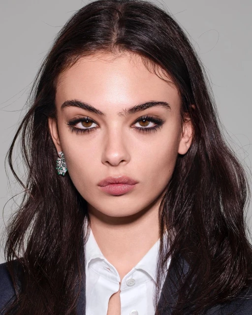 Close-up portrait of a young woman with brown hair, wearing a blazer, earring, and makeup against a gray background.