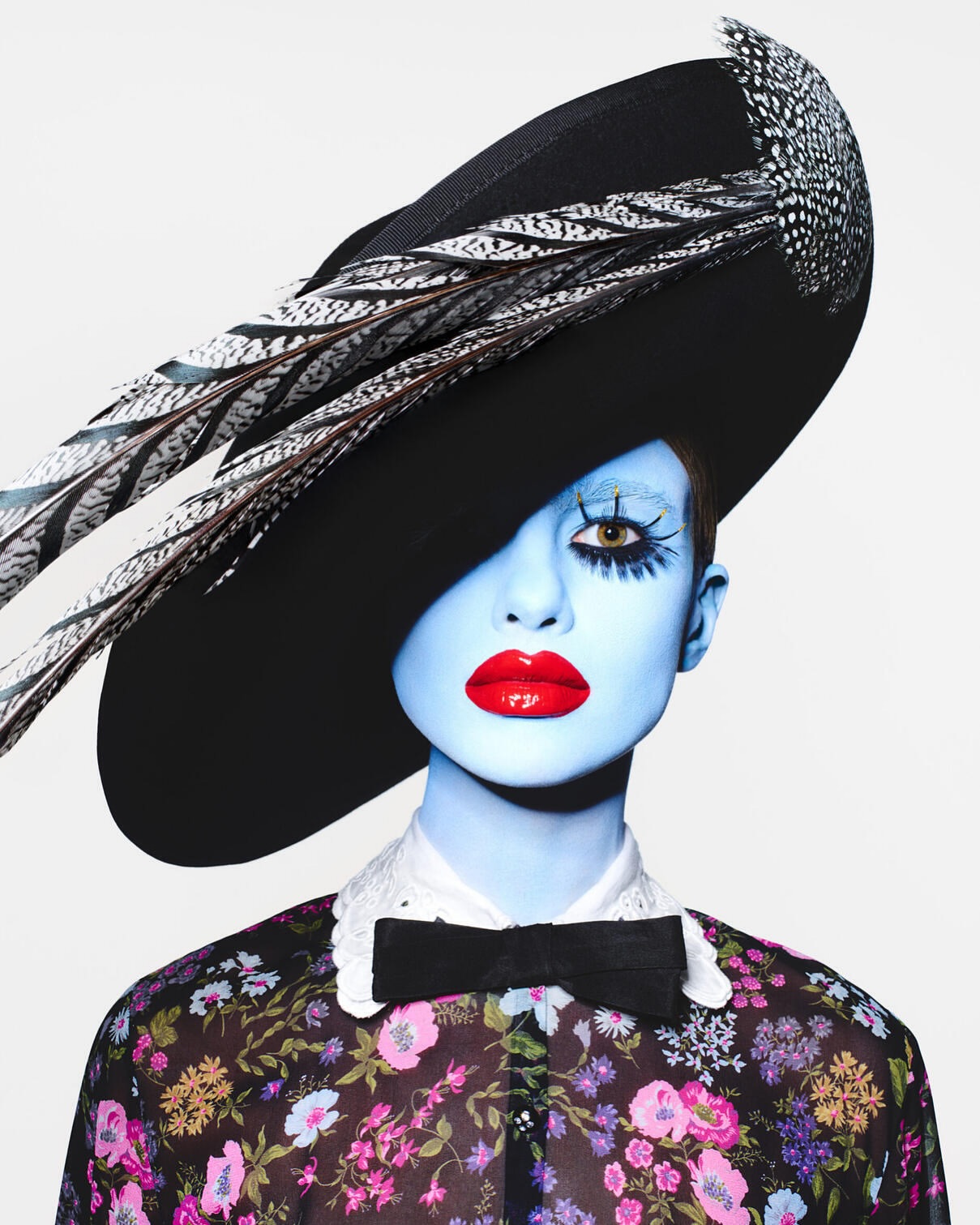 Fashion portrait of a model with pale blue skin, striking red lips, and a black hat adorned with feathers against a white backdrop.