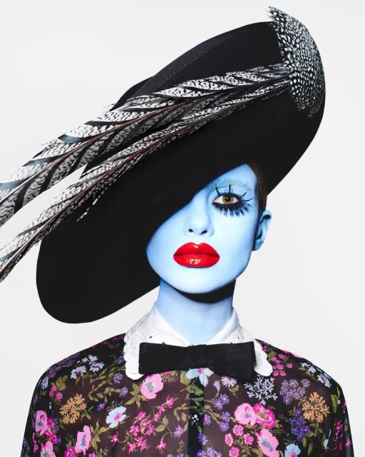 Fashion portrait of a model with pale blue skin, striking red lips, and a black hat adorned with feathers against a white backdrop.