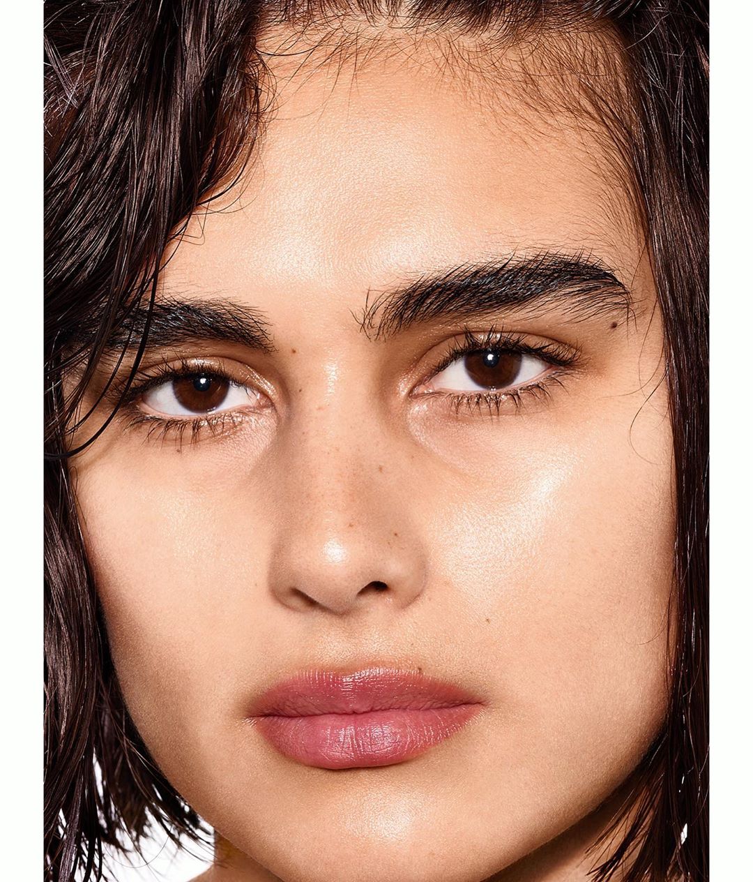 Close-up portrait of a young adult female with brown eyes and wet, dark brown hair, styled with a subtle pink lip colour and clear facial skin.