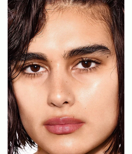 Close-up portrait of a young adult female with brown eyes and wet, dark brown hair, styled with a subtle pink lip colour and clear facial skin.