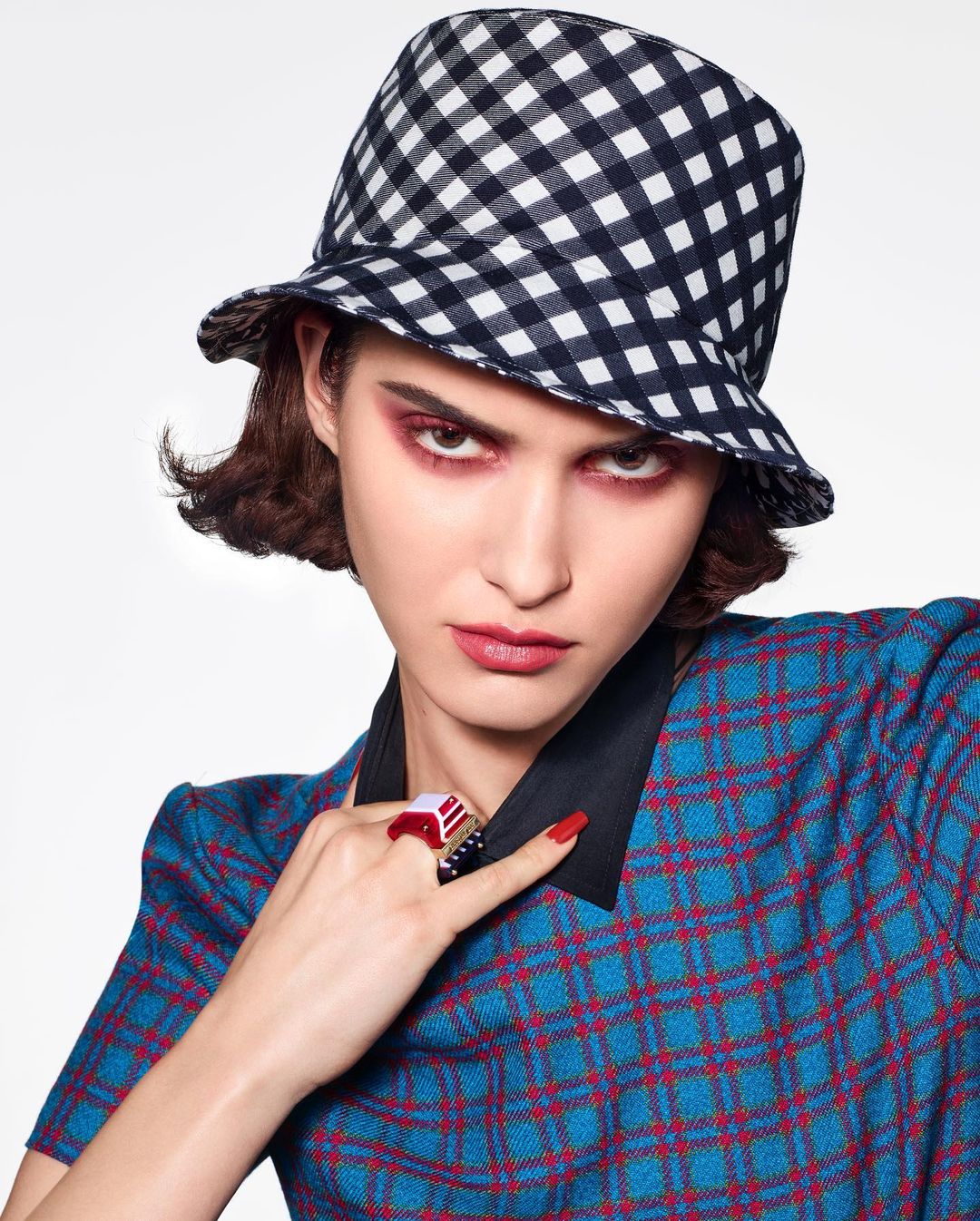 Fashion portrait of woman wearing a blue and red plaid dress, black and white checkered bucket hat, and statement ring against white background.