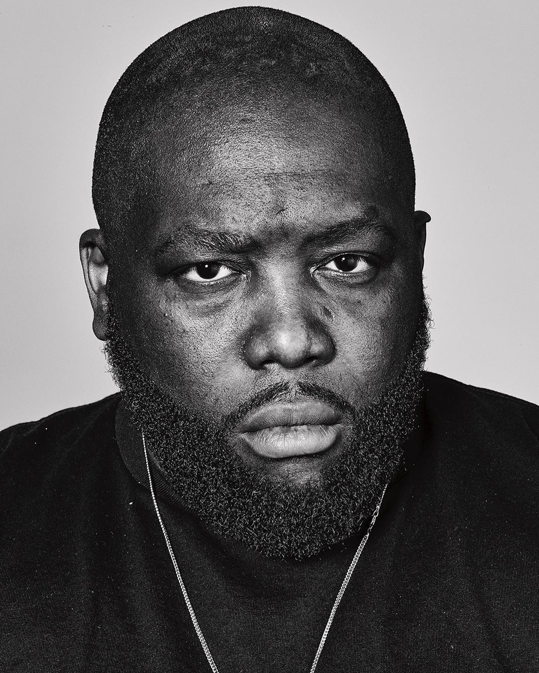 Monochrome headshot of a Black male with a beard, wearing a black shirt and a silver chain, against a plain grey backdrop.
