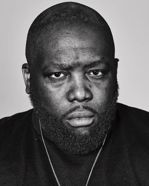 Monochrome headshot of a Black male with a beard, wearing a black shirt and a silver chain, against a plain grey backdrop.