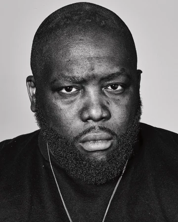 Monochrome headshot of a Black male with a beard, wearing a black shirt and a silver chain, against a plain grey backdrop.