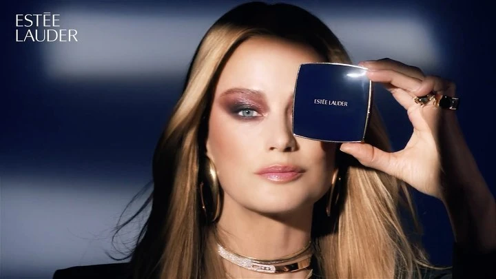 Blonde woman with gold jewelry poses indoors holding Estée Lauder compact, wearing dramatic makeup and a black jacket in a beauty shot.