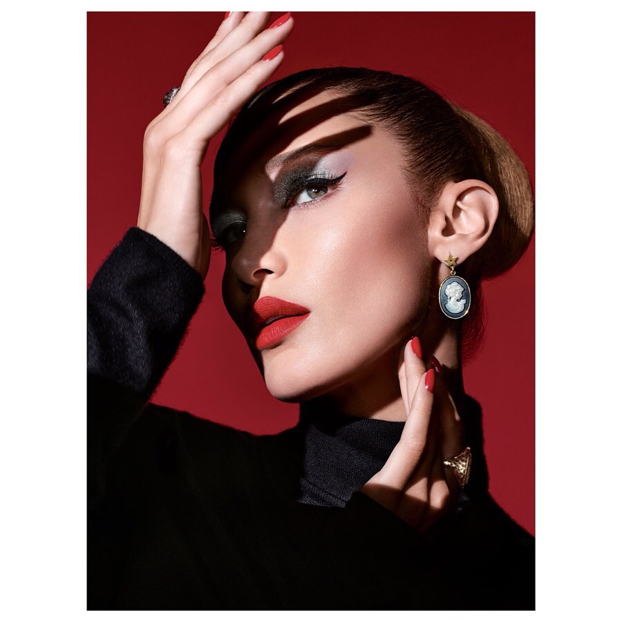 Close-up beauty shot of a woman with red lips, smokey eye makeup, and black turtleneck, accessorized with statement earrings on a red background.