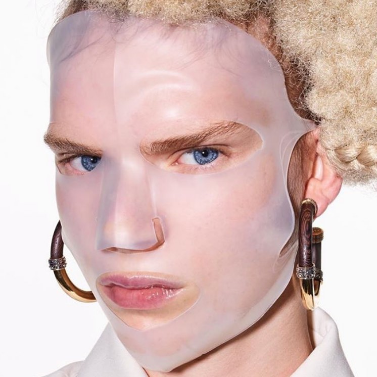 Close-up of a person with blue eyes wearing a translucent silicone face mask and large hoop earrings against a white background.