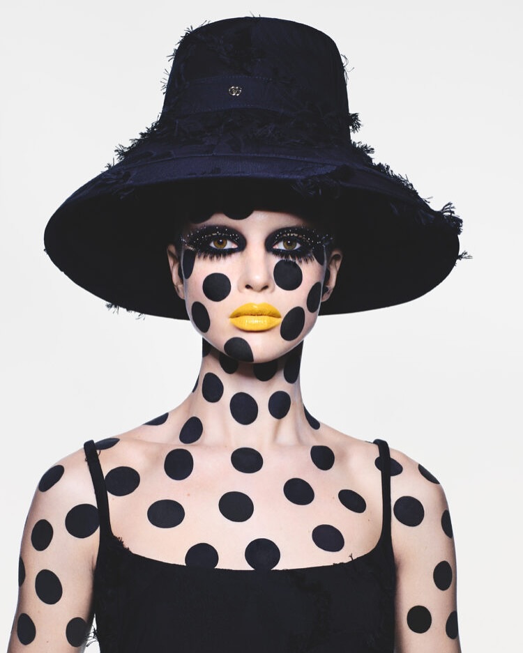 A woman wearing a black hat and black polka dots on her skin, with yellow lips and large eyelashes, poses against a white background.