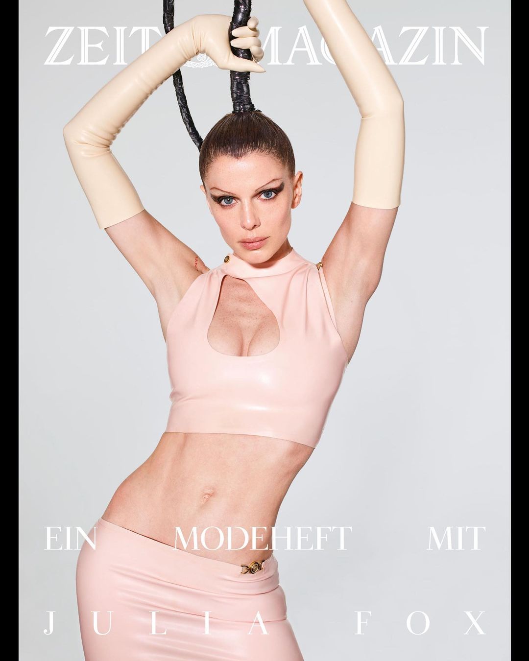 Julia Fox poses in a glossy pale pink two-piece outfit with matching gloves, set against a light gray backdrop for a magazine cover.