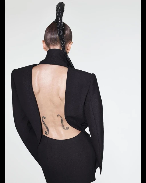 Adult female with avant-garde braided updo, black open-back dress, and back tattoos posing in a bright white photo studio.
