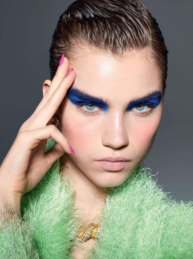 Close-up portrait of a young adult woman with striking blue eyeshadow, glossy hair, pink nails, and a fuzzy green garment, against a gray backdrop.