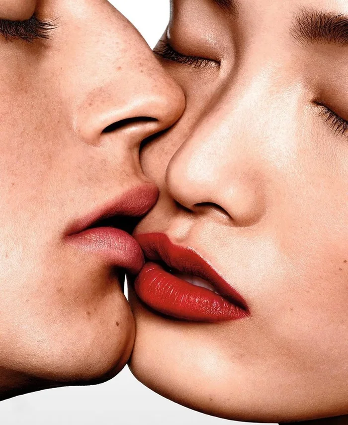 Close-up portrait of two models kissing, featuring vibrant red lipstick against a clean white background by Richard Burbridge.
