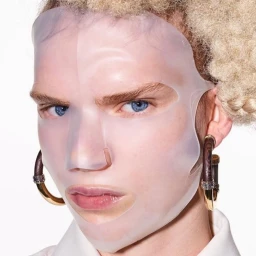 Close-up portrait of a person wearing a translucent face mask and large hoop earrings against a white background