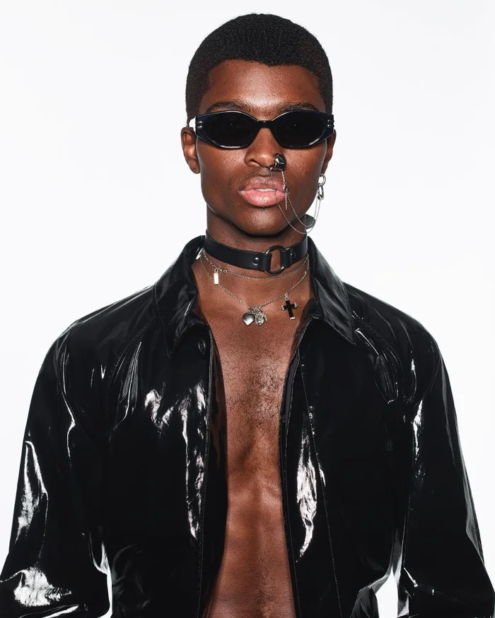 Black man wearing dark sunglasses, septum chain, choker, and open black vinyl shirt against white background.