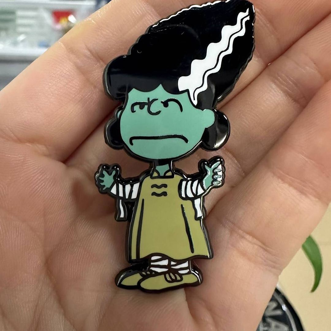 Enamel pin of cartoon woman with green skin, black hair, and a pale green dress, held in a person's hand with blurred background.