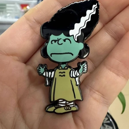 Enamel pin of cartoon woman with green skin, black hair, and a pale green dress, held in a person's hand with blurred background.
