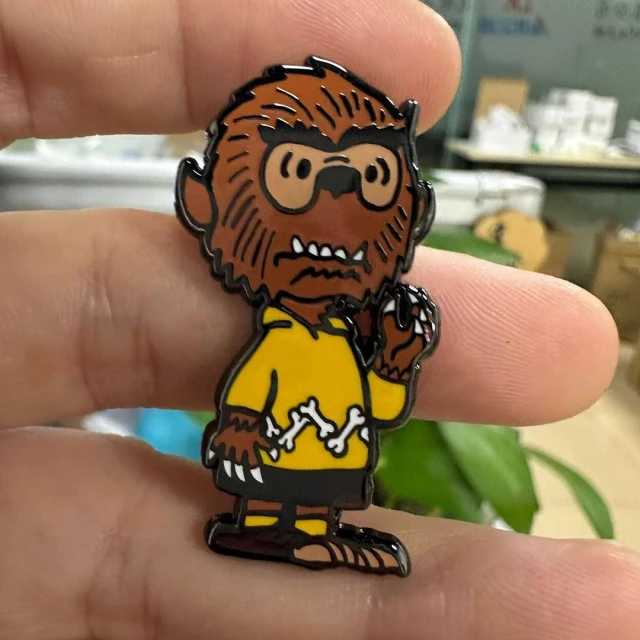 Cartoon werewolf character pin.  Character is wearing glasses and a yellow shirt. Pin is held by a human hand.