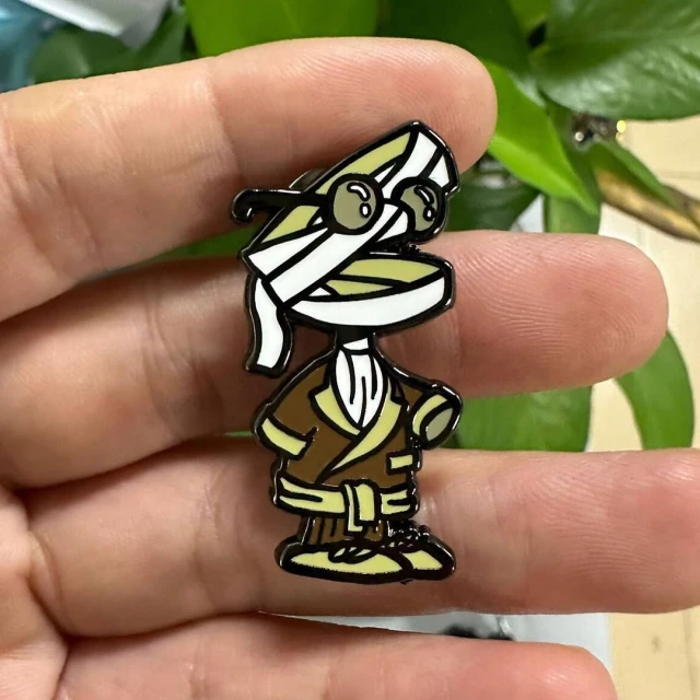 Enamel pin of a cartoon mummy wearing round glasses and a robe on a neutral background, held in a person's hand.