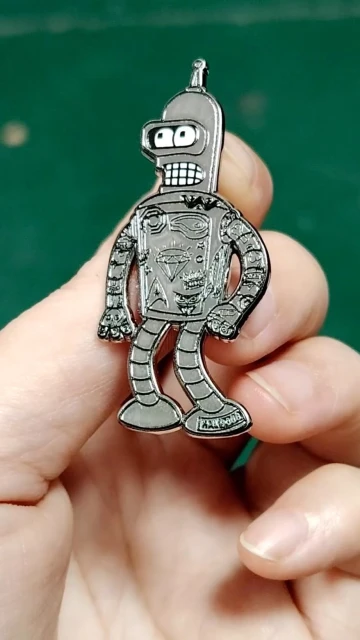 Robot cartoon character Bender pin is held in a person's hand against a green background in a close-up studio shot.