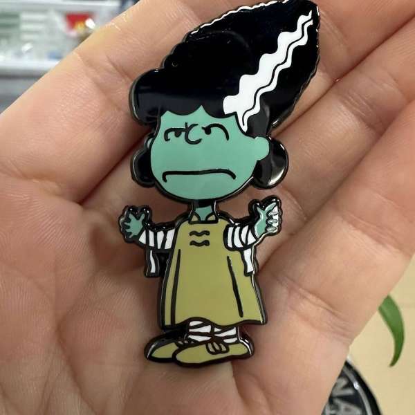 Close-up of a cartoon character pin with a green face, black hair, and an olive-green outfit, held in a person's hand.