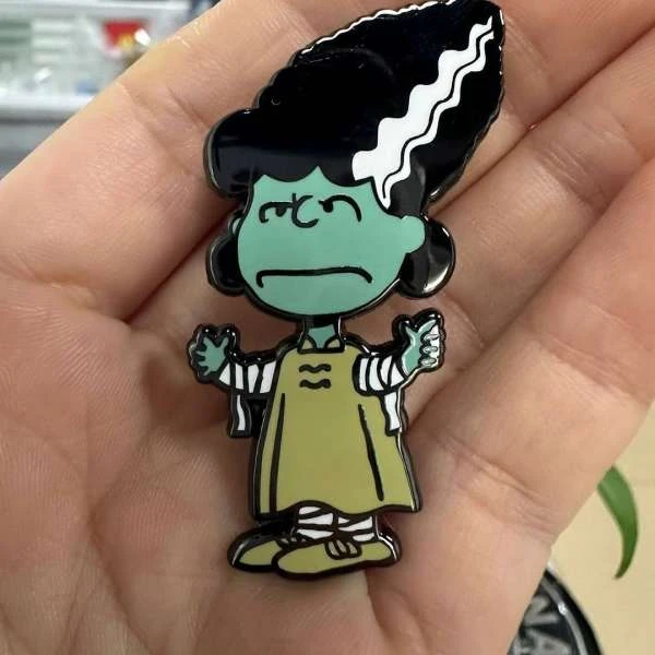 Close-up of a cartoon character pin with a green face, black hair, and an olive-green outfit, held in a person's hand.