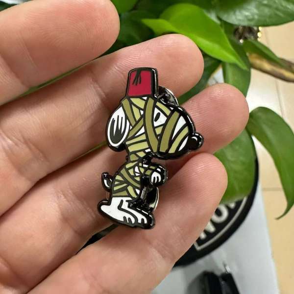 Close-up of Snoopy mummy enamel pin held in a person's hand with blurred green plant in the background.
