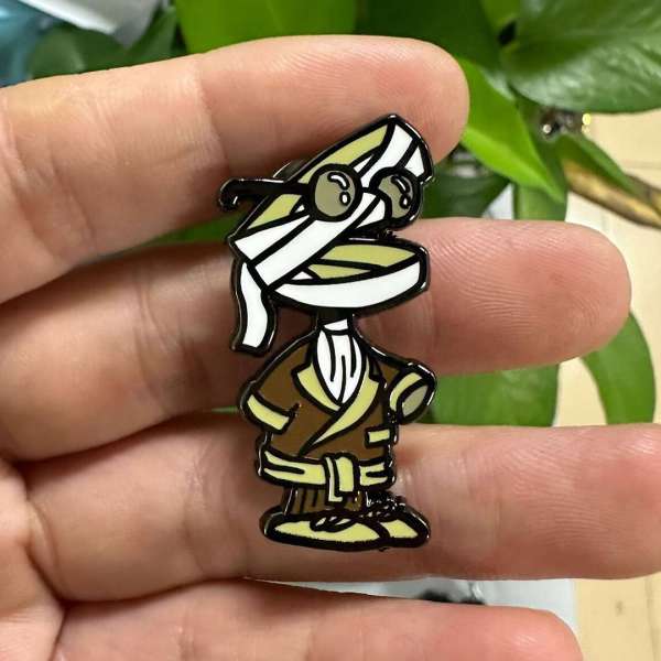 Cartoon mummy character pin with round glasses and brown robe, held in hand, with blurred green plant in the background.