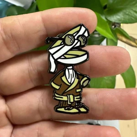 Cartoon mummy character pin with round glasses and brown robe, held in hand, with blurred green plant in the background.