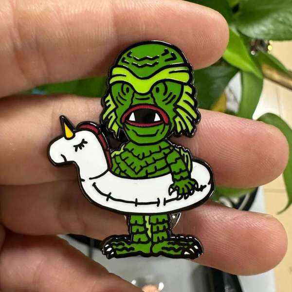 Creature from the Black Lagoon pin wearing a unicorn floatie, held in a person's hand with blurred plants and surfaces in the background.