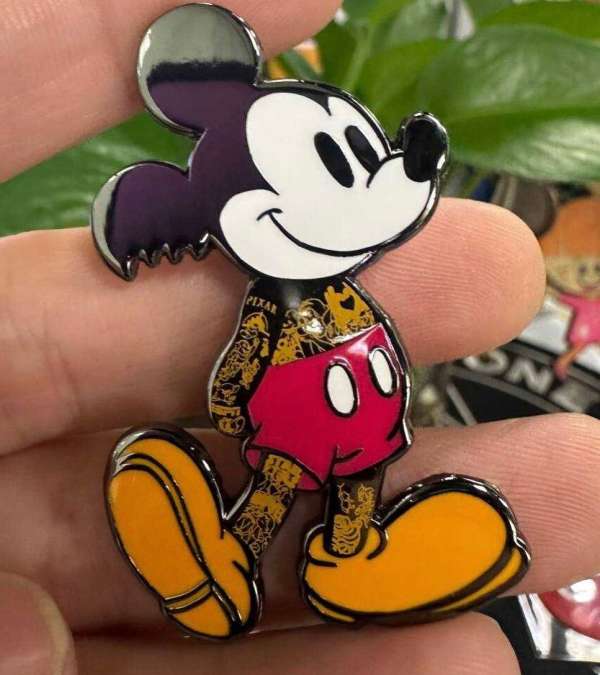 Close-up of a Mickey Mouse pin, featuring a colorful, tattooed version of the cartoon character, held in a person's hand.