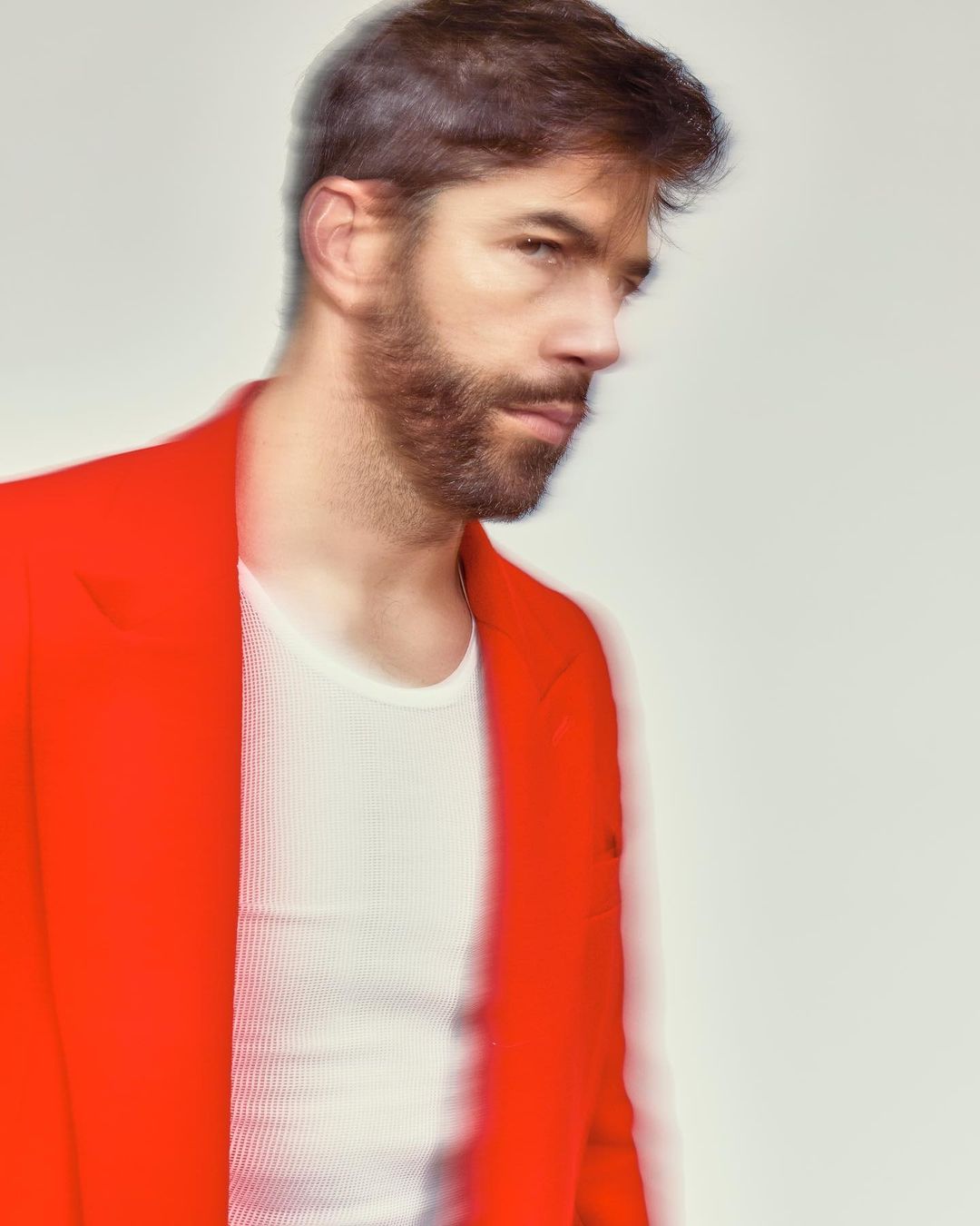 Man in red blazer and white ribbed t-shirt poses in a close-up indoor shot on a white background with soft lighting and a shallow depth of field.