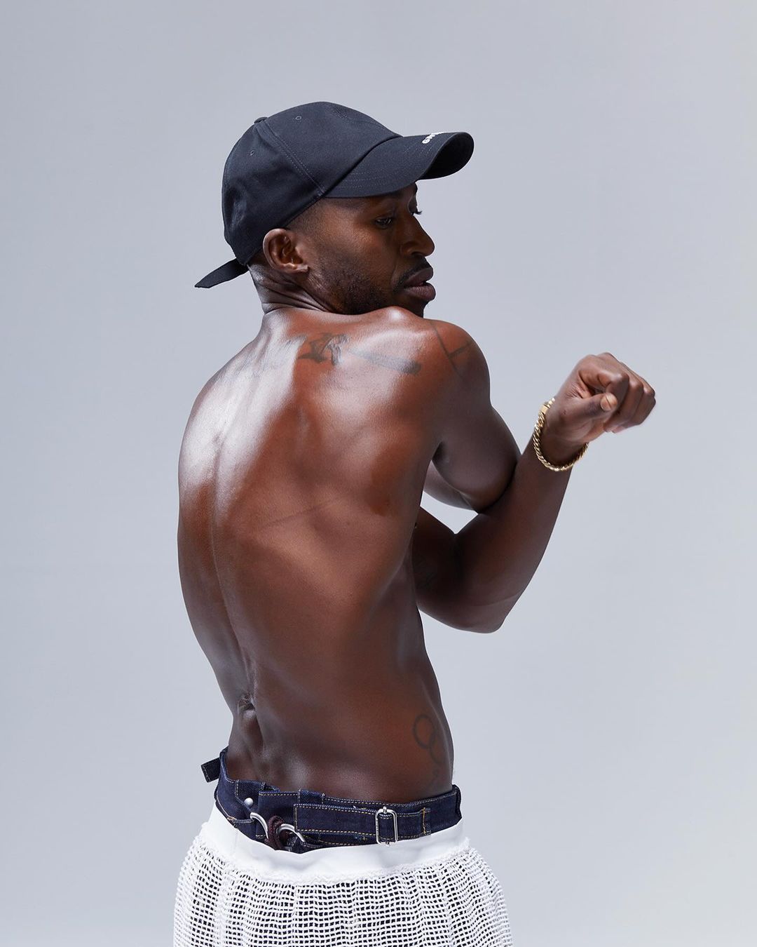 Portrait of a bare-chested African-American man in a cap with a gold bracelet, white skirt, and denim belt standing in a photo studio.