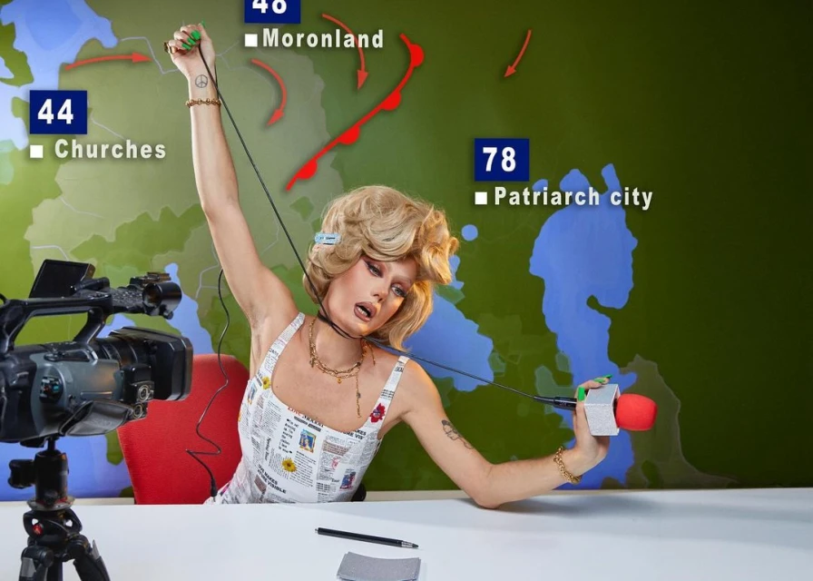 Adult female in news-print dress with dramatic makeup sits at desk with microphone, camera, and monitor displaying map of the world.