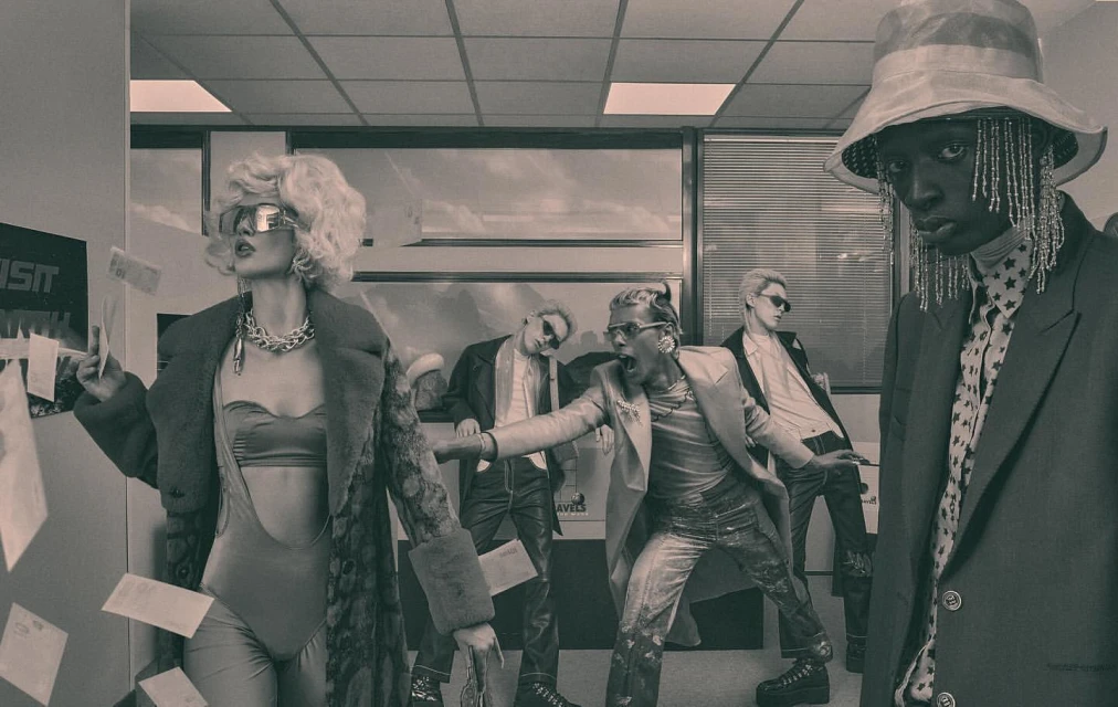 Editorial fashion shot featuring five fashionably dressed adults in an office setting with papers falling, set in monochrome.