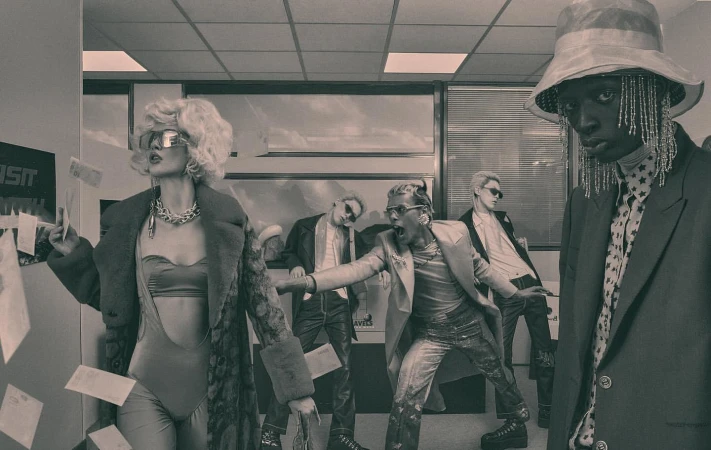Editorial fashion shot featuring five fashionably dressed adults in an office setting with papers falling, set in monochrome.